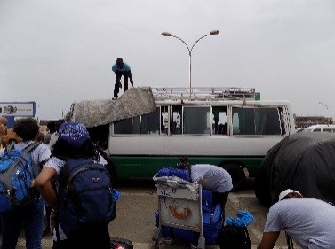 Drexel students, faculty and staff wearing backpacks stand in a parking lot in Senegal while a man covers a buss with a tarp.